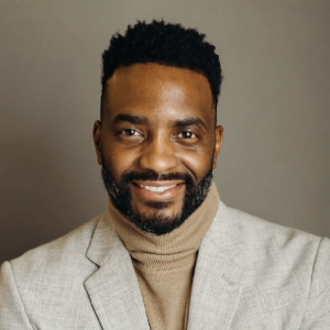 Sedric Brinson Smiling person with short dark hair and full facial hair wearing a cream turtleneck and a cream blazer.