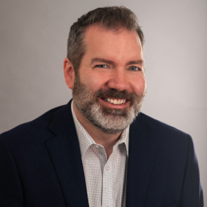 Christopher Morris Smiling person with salt and pepper short hair and beard wearing a suit jacket and a light colored shirt with small grid pattern on it