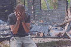 disaster Person covered in mud sitting with hands covering their face with rubble behind them.