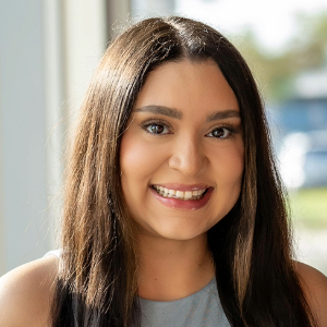 Angelica_Bravo Altra Smiling person with long brown hair parted in the middle wearing a pink lipstick and a gray tank style top