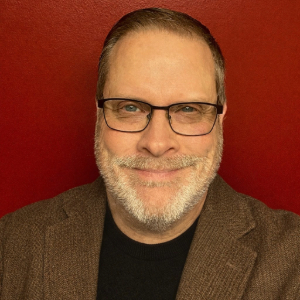 Phillip Crocker Person with short brown hair and white facial hair wearing glasses and a brown blazer over a black shirt